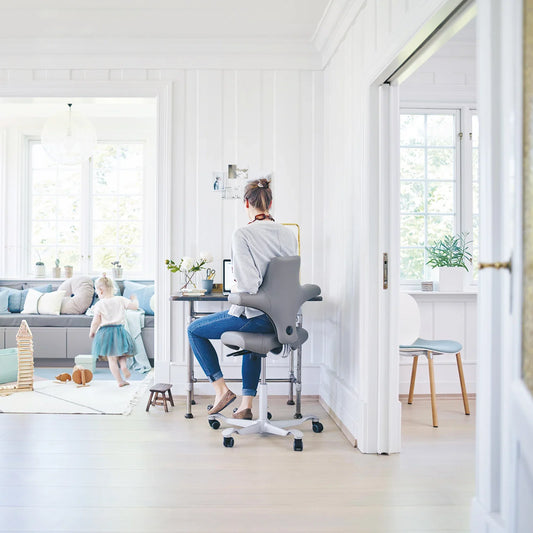 Vrouw werkt aan een bureau in de woonkamer op een zadelvormige bureaustoel, met een spelend kind op de achtergrond